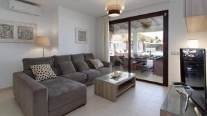 Ein modernes Wohnzimmer mit einem grauen Sofa, einem Glastisch und einer Schiebetür zu einem Außenbereich mit Esstisch und Stühlen.