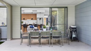 Eine moderne Terrasse mit Esstisch und Stühlen, einem Grill und einem durch eine Glastür einsehbaren Küchenbereich.