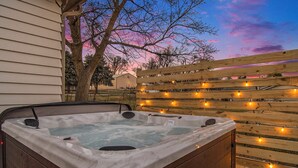 Un jacuzzi con cubierta de madera y luces colgantes, frente a una casa y un árbol.