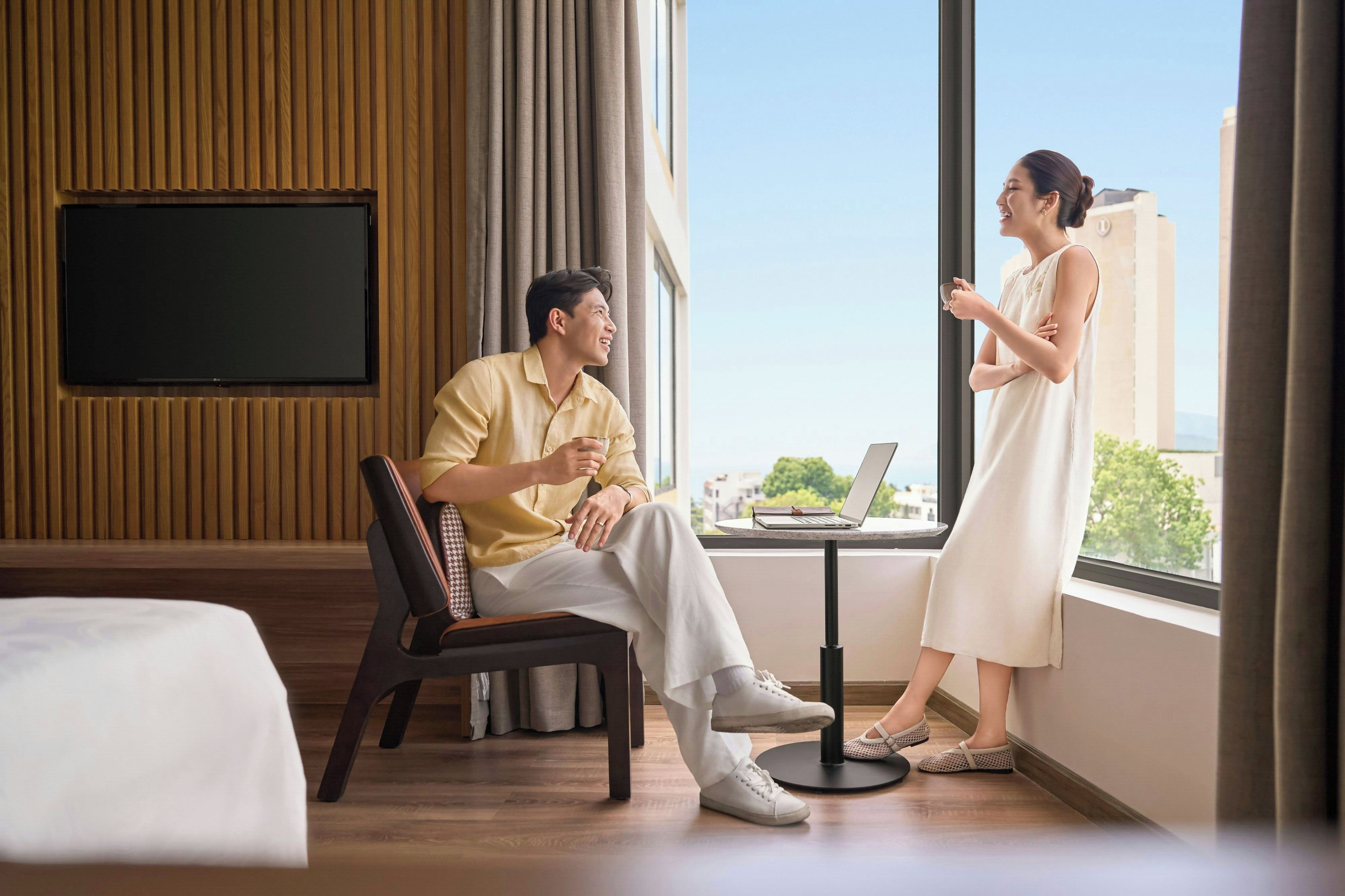 A modern hotel room with a large window, a flat-screen TV mounted on the wall, a small round table with a laptop, and a comfortable seating area with a chair and a sofa.