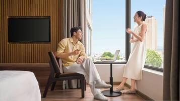 A modern hotel room with a large window, a flat-screen TV mounted on the wall, a small round table with a laptop, and a comfortable seating area with a chair and a sofa.
