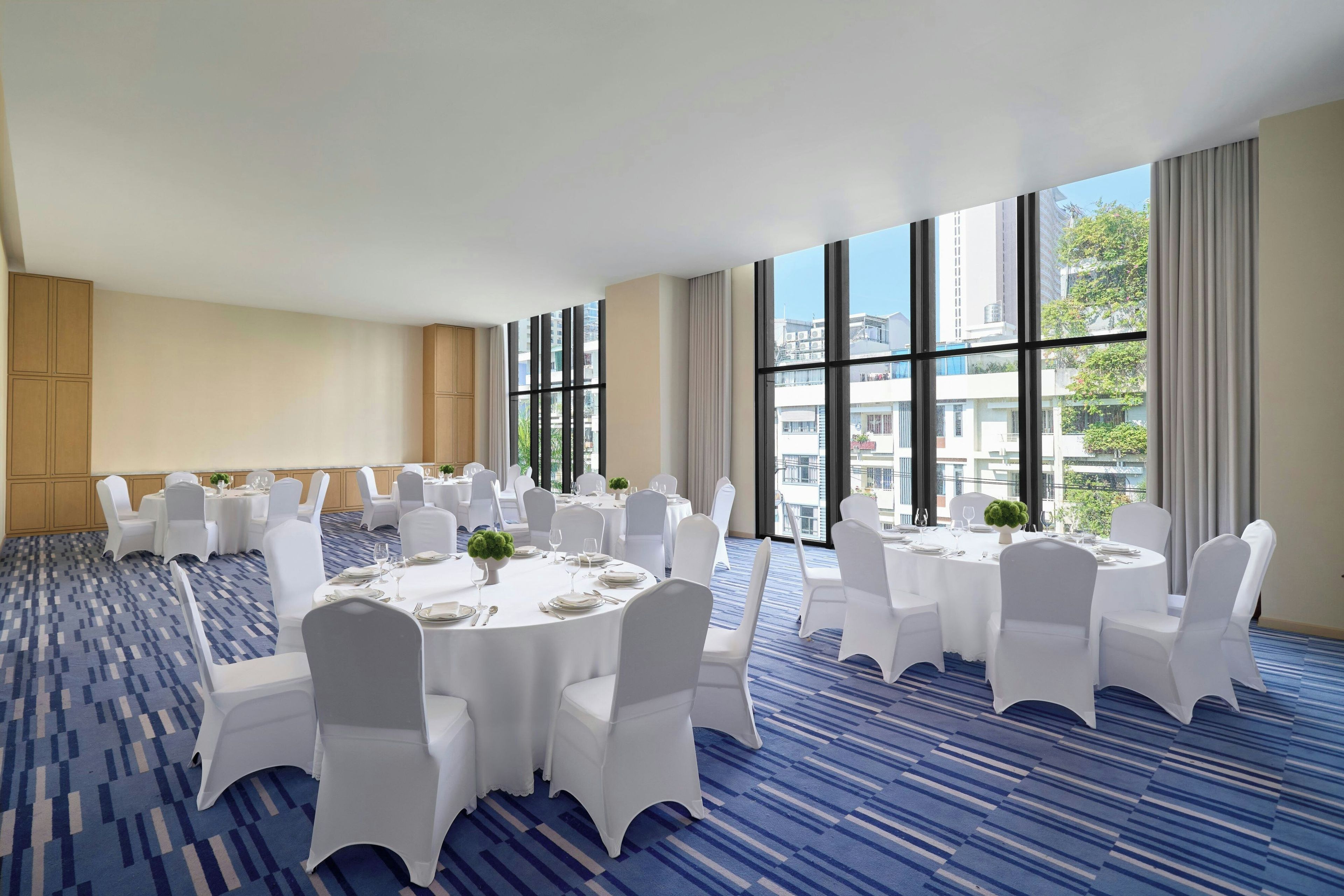 Un salón de conferencias con mesas redondas, sillas blancas y vistas a edificios a través de amplias ventanas.