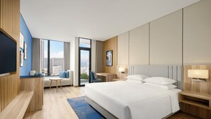 Ein Hotelzimmer mit einem großen Bett, einem Schreibtisch, einem Sessel, einem Fernseher und Blick auf die Stadt.