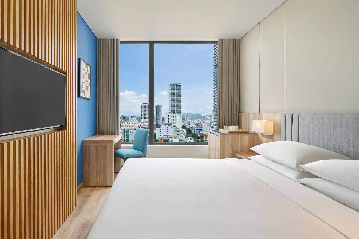 Une chambre d’hôtel moderne dotée d’un grand lit, d’un bureau et d’une chaise, et offrant une vue sur la ville grâce à de grandes fenêtres.