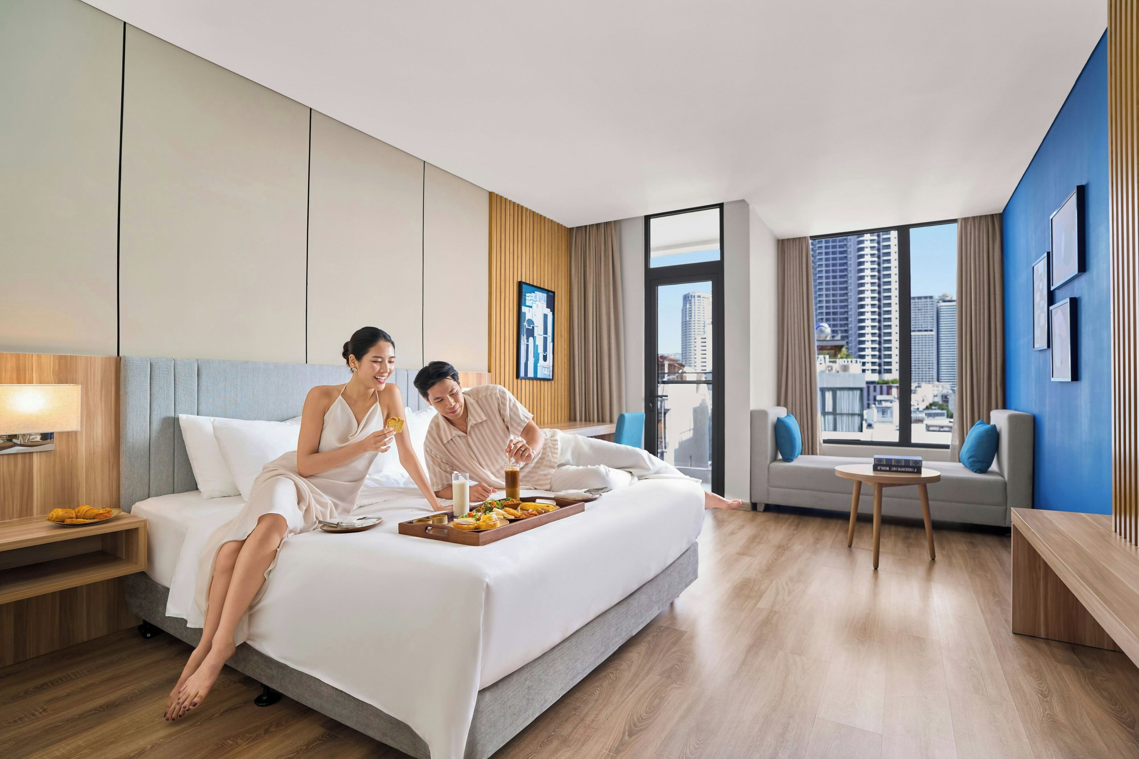 A modern hotel room with a large bed, a small sofa, a side table, and a view of the city through large windows.