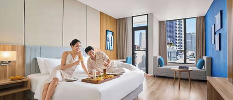 A modern hotel room with a large bed, a small sofa, a side table, and a view of the city through large windows.