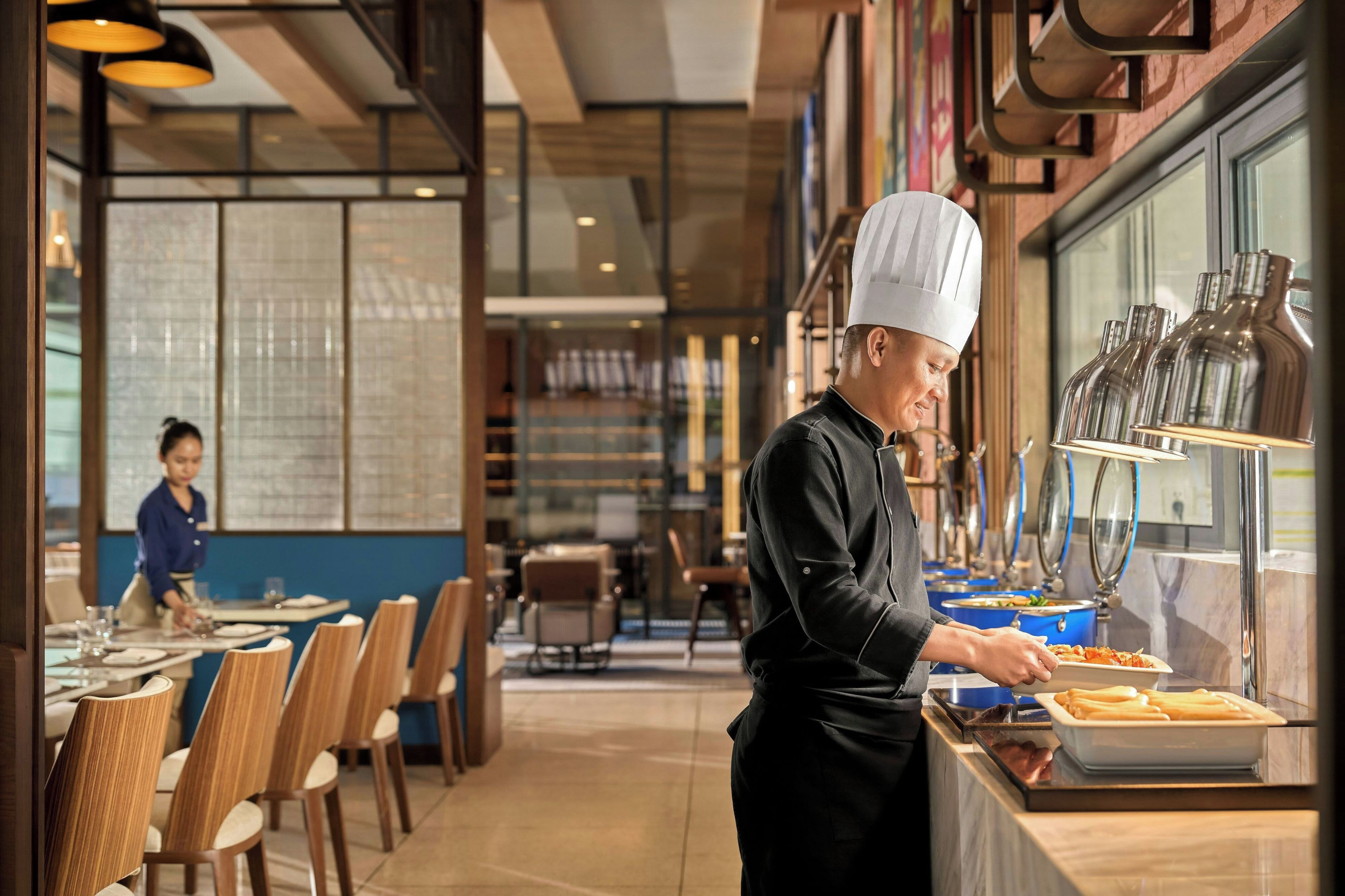 A restaurant interior with a chef preparing food at a counter, tables with chairs, and a buffet area with food containers.