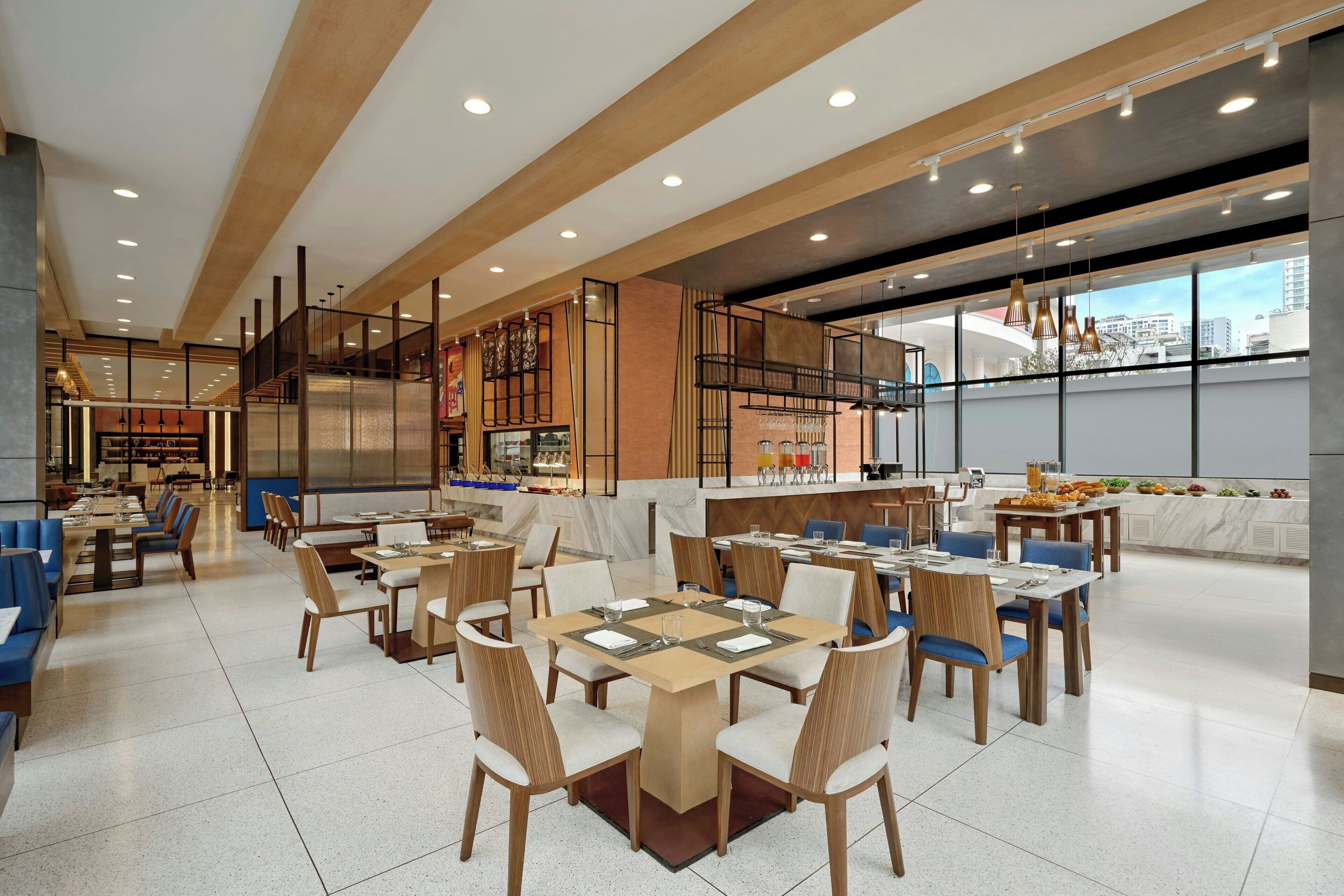 A modern dining area with multiple tables and chairs, a kitchen area with a counter, and large windows offering a view of the city.