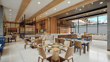 A modern dining area with multiple tables and chairs, a kitchen area with a counter, and large windows offering a view of the city.