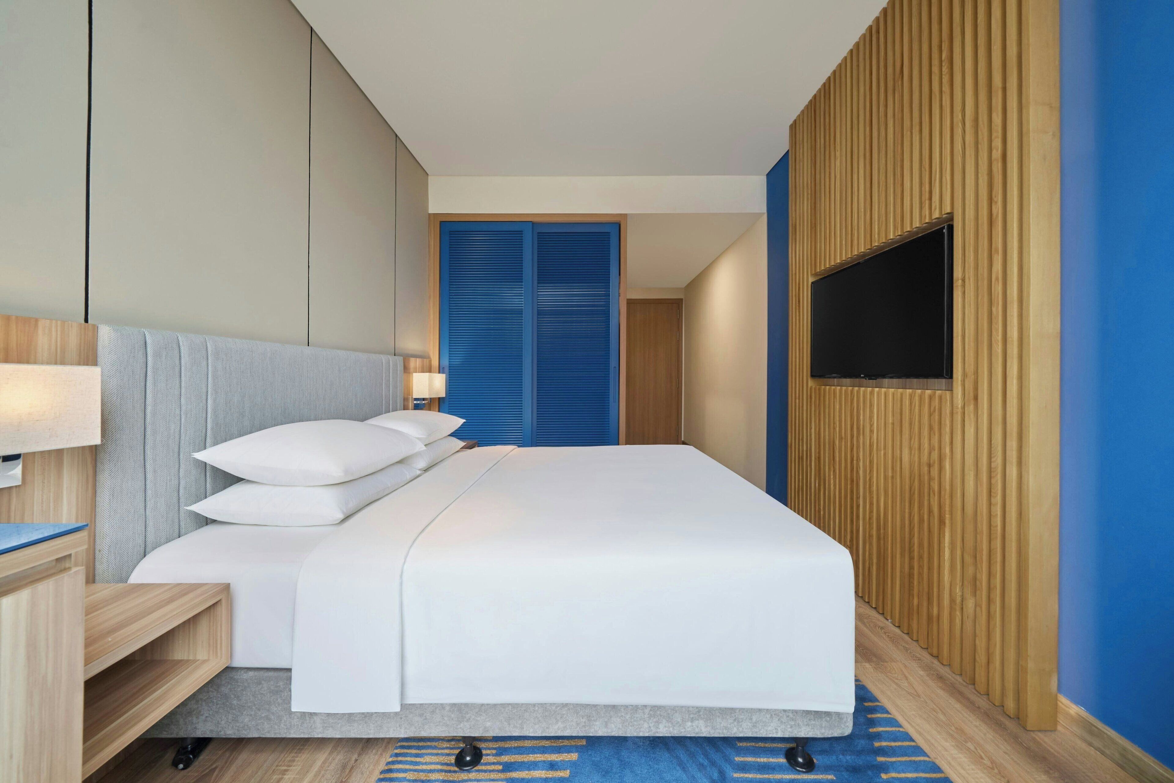 A modern hotel room with a bed, bedside table, lamp, and a TV mounted on the wall.