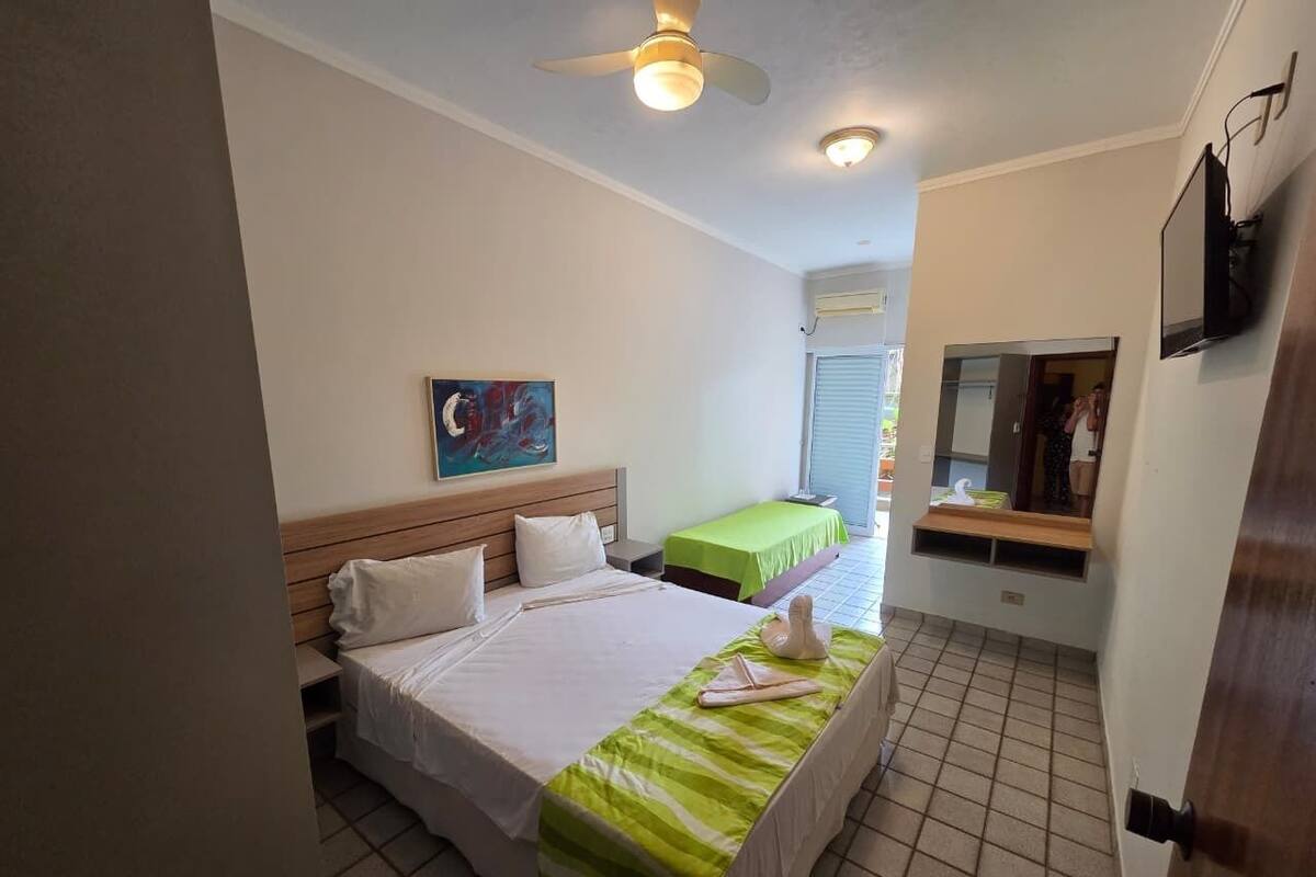 A hotel room with a large bed, a smaller bed, a painting on the wall, a ceiling fan, and a TV mounted on the wall.
