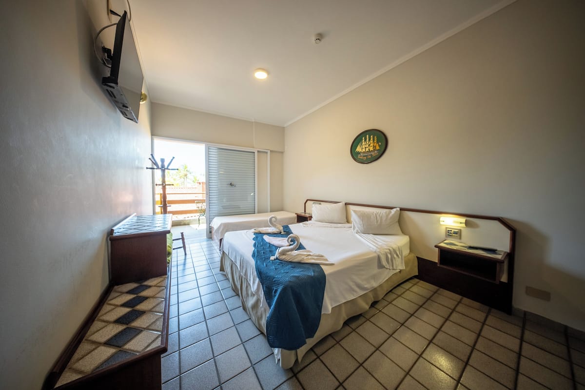 A hotel room with two beds, a small desk, a wall clock, and a window with blinds.