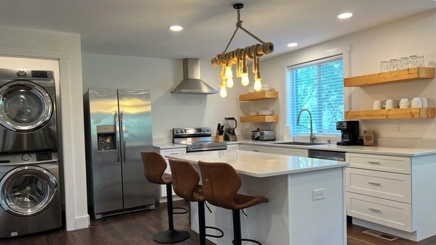 Una cocina moderna con una isla central, taburetes de bar, lavadora y secadora integradas, y estanterías de madera.