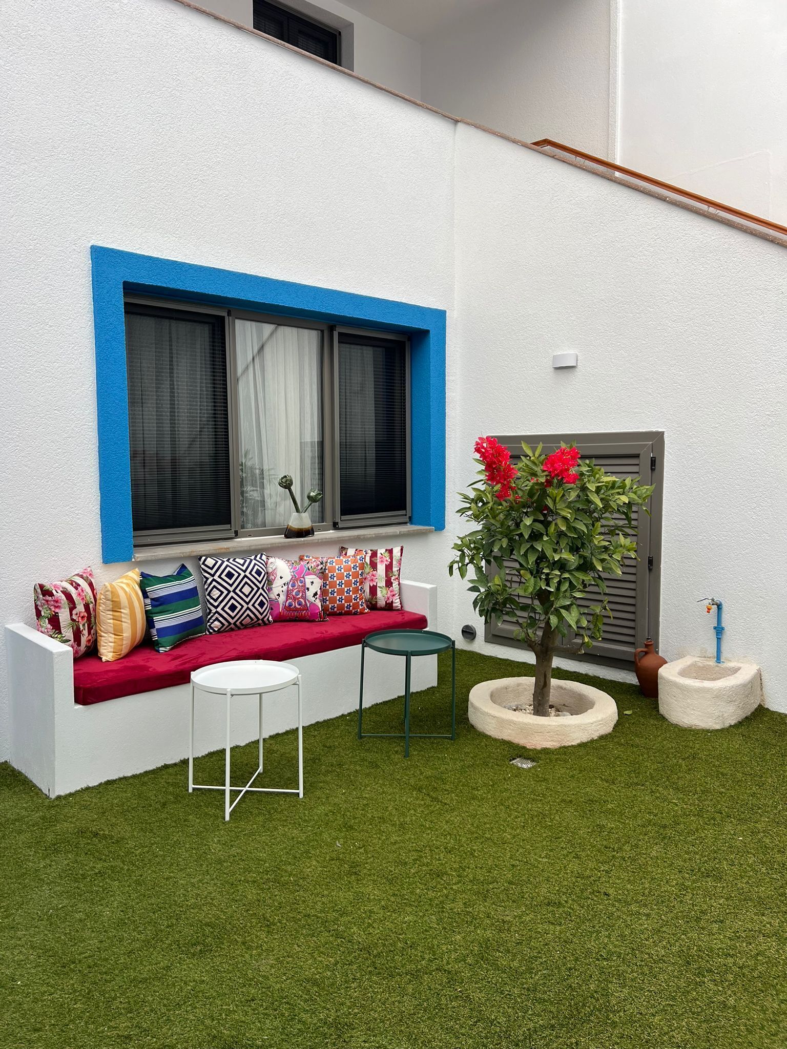 A modern outdoor seating area with a white wall, blue trim, and a red bench. There are two white tables and a potted plant with red flowers.