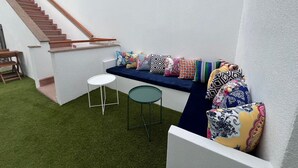 A small, enclosed outdoor space with a wooden staircase, a blue cushioned bench with colorful cushions, and a small round table.