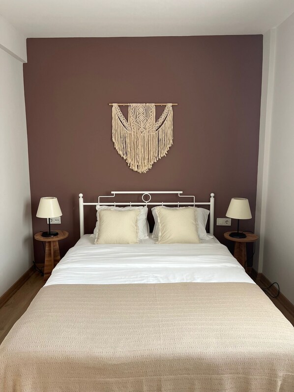 Una cama bien hecha con una colcha blanca y almohadas, un colgador de macramé en la pared y dos lámparas de noche sobre mesitas de madera.