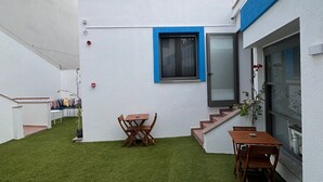 A modern building with a flat roof, white exterior walls, and blue window frames. There is a small outdoor seating area with a wooden table and chairs.