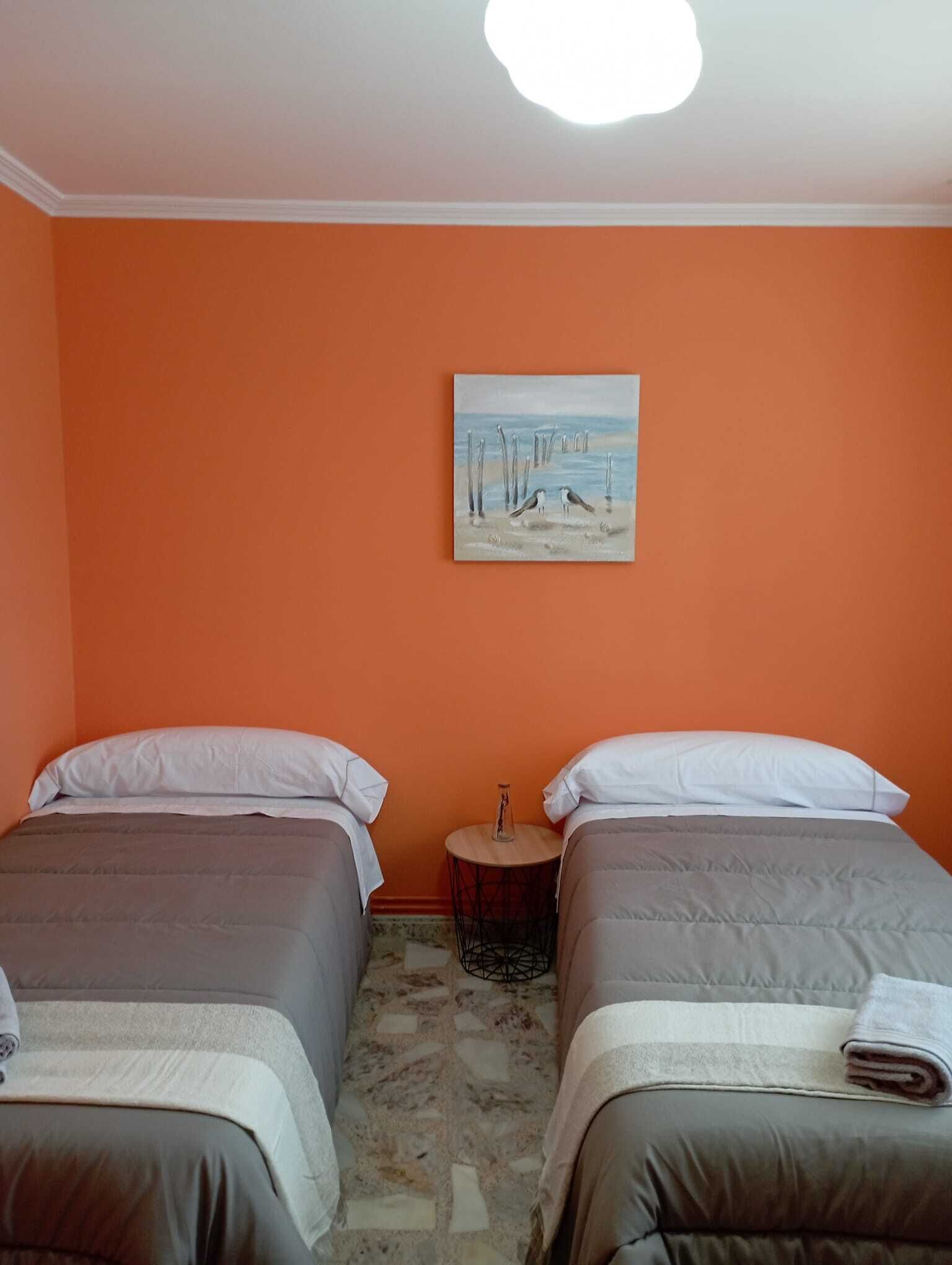 Una habitación con dos camas, paredes anaranjadas, un cuadro de pájaros y una mesita redonda.