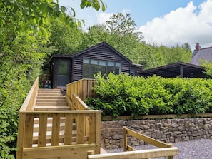 A wooden staircase leading to a dark wooden building with a stone wall and greenery around. - Lodge at Nadur (Machynlleth)