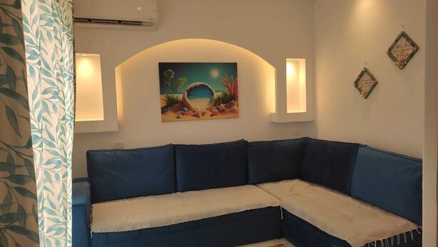 Sala de estar com canto, sofá modular azul, tapete listrado e colorido, e pintura com tema praiano na parede.