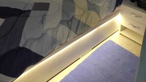 Cama bem arrumada com um cobertor listrado e um brinquedo de pelúcia. Há uma mesinha de cabeceira com uma luminária e uma gaveta pequena.