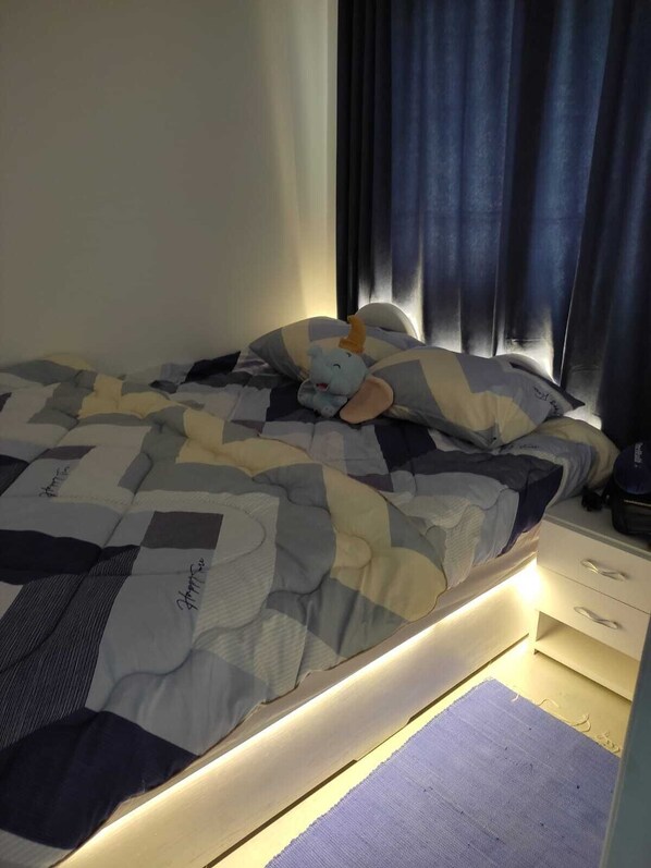 A neatly made bed with a striped blanket and a plush toy. There is a bedside table with a lamp and a small drawer.