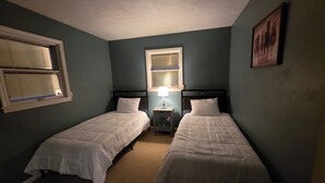 Un dormitorio con dos camas individuales, una mesita de noche con lámpara, una ventana con persianas y un cuadro en la pared.