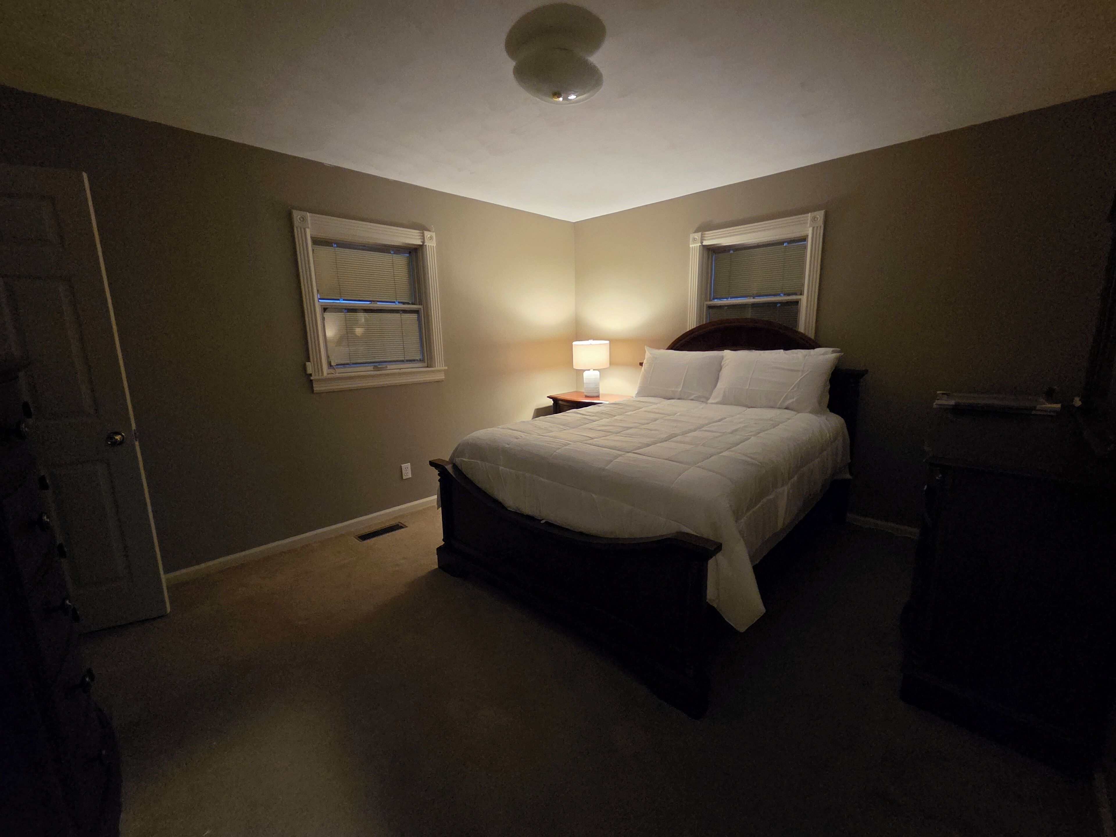 Une chambre à coucher avec un lit, deux fenêtres équipées de stores, une table de chevet avec une lampe et un ventilateur de plafond.