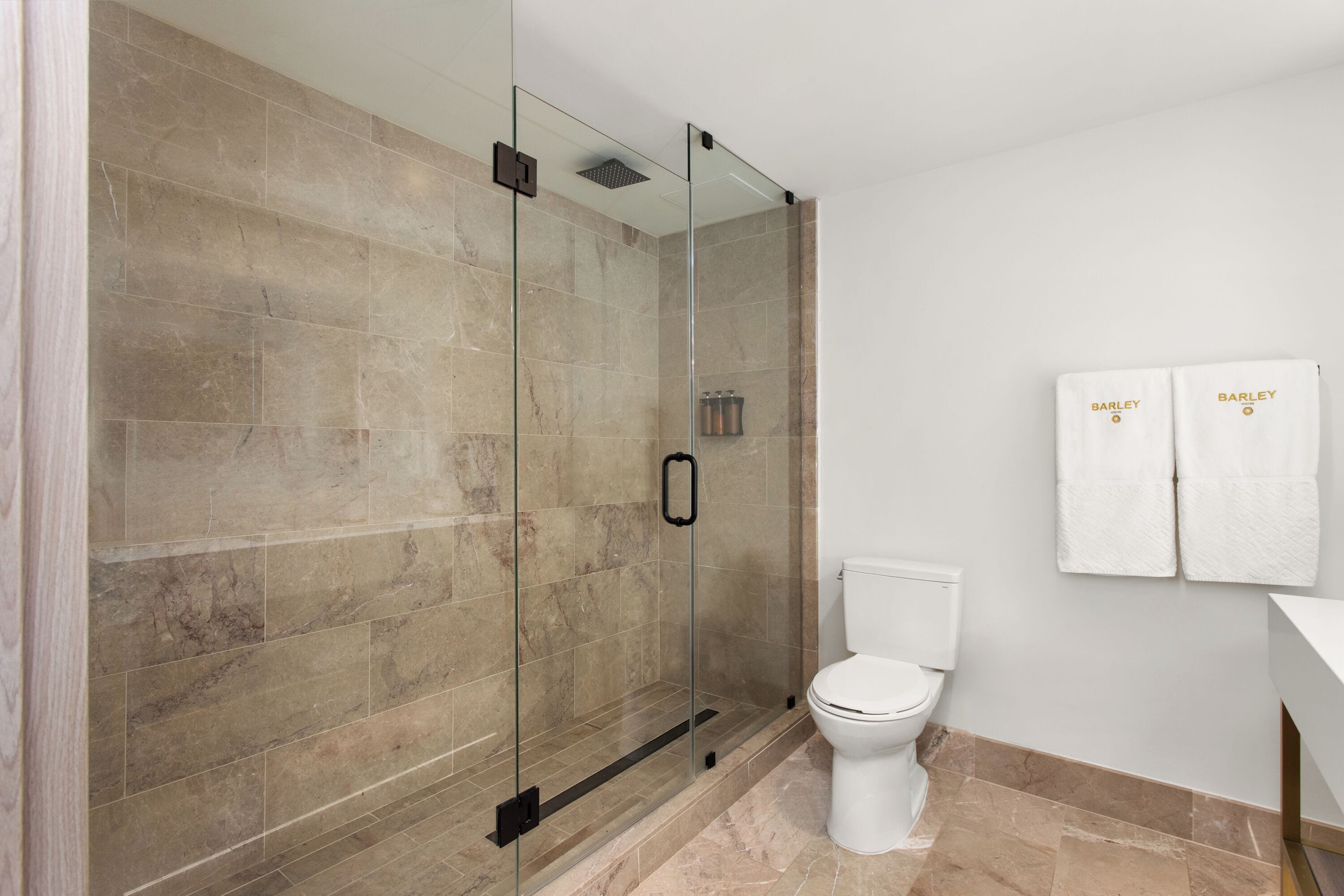 A modern bathroom with a glass-enclosed shower, a white toilet, and two towels hanging on the wall.