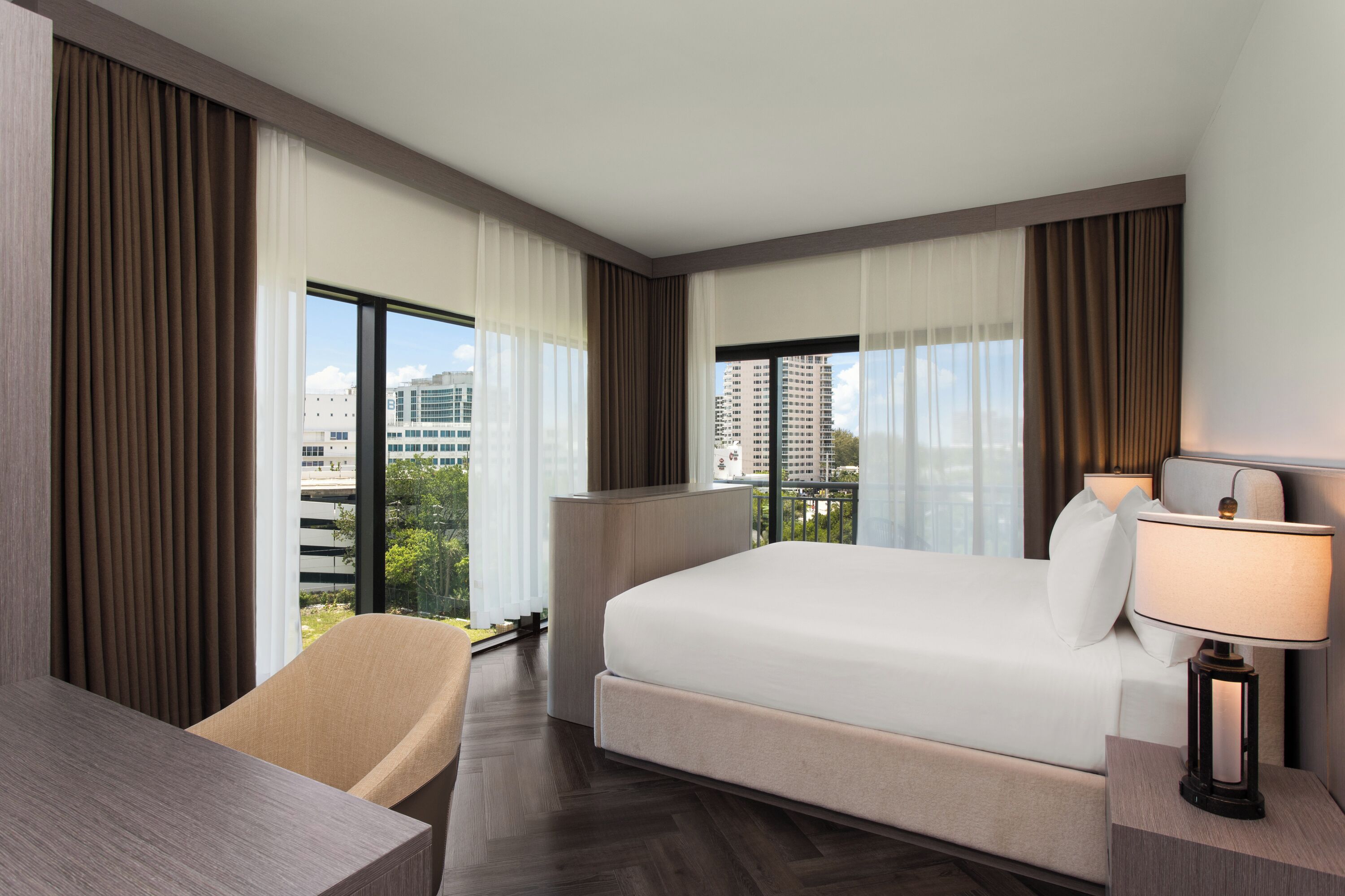 Una habitación de hotel con cama, escritorio, silla, lámpara y ventanas grandes con cortinas.