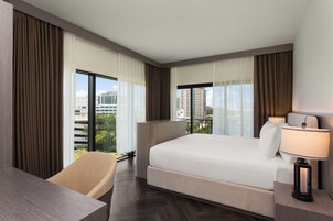 Quarto de hotel com cama, escrivaninha, cadeira, luminária e janelas amplas com cortinas.