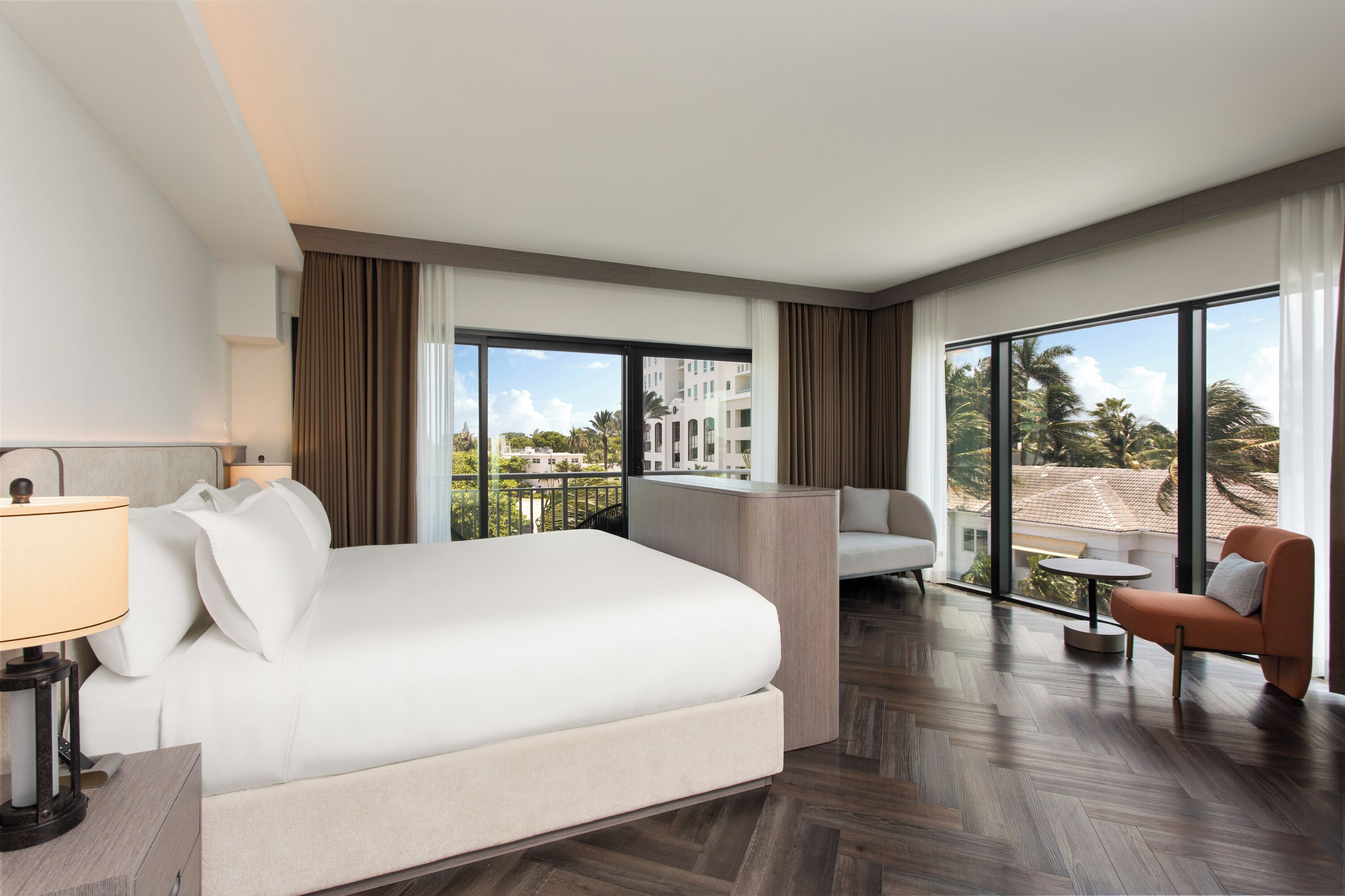 A hotel room with a large bed, a desk, a chair, and a view of the cityscape through large windows.