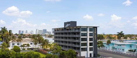 A modern multi-story hotel with a pool area, surrounded by greenery and a city skyline in the background.