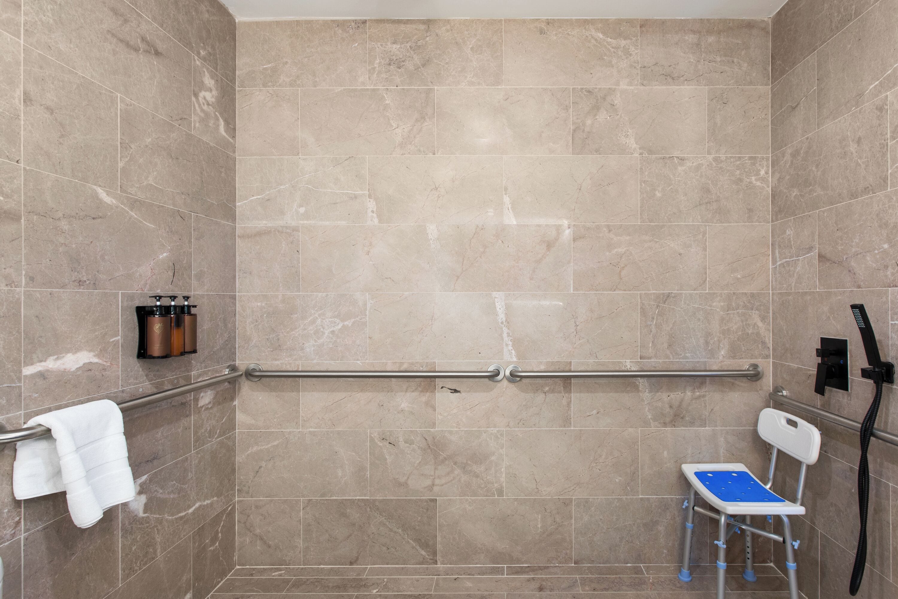 Zona de ducha con azulejos color beige, una barra de apoyo, una toalla blanca y un asiento de ducha azul.