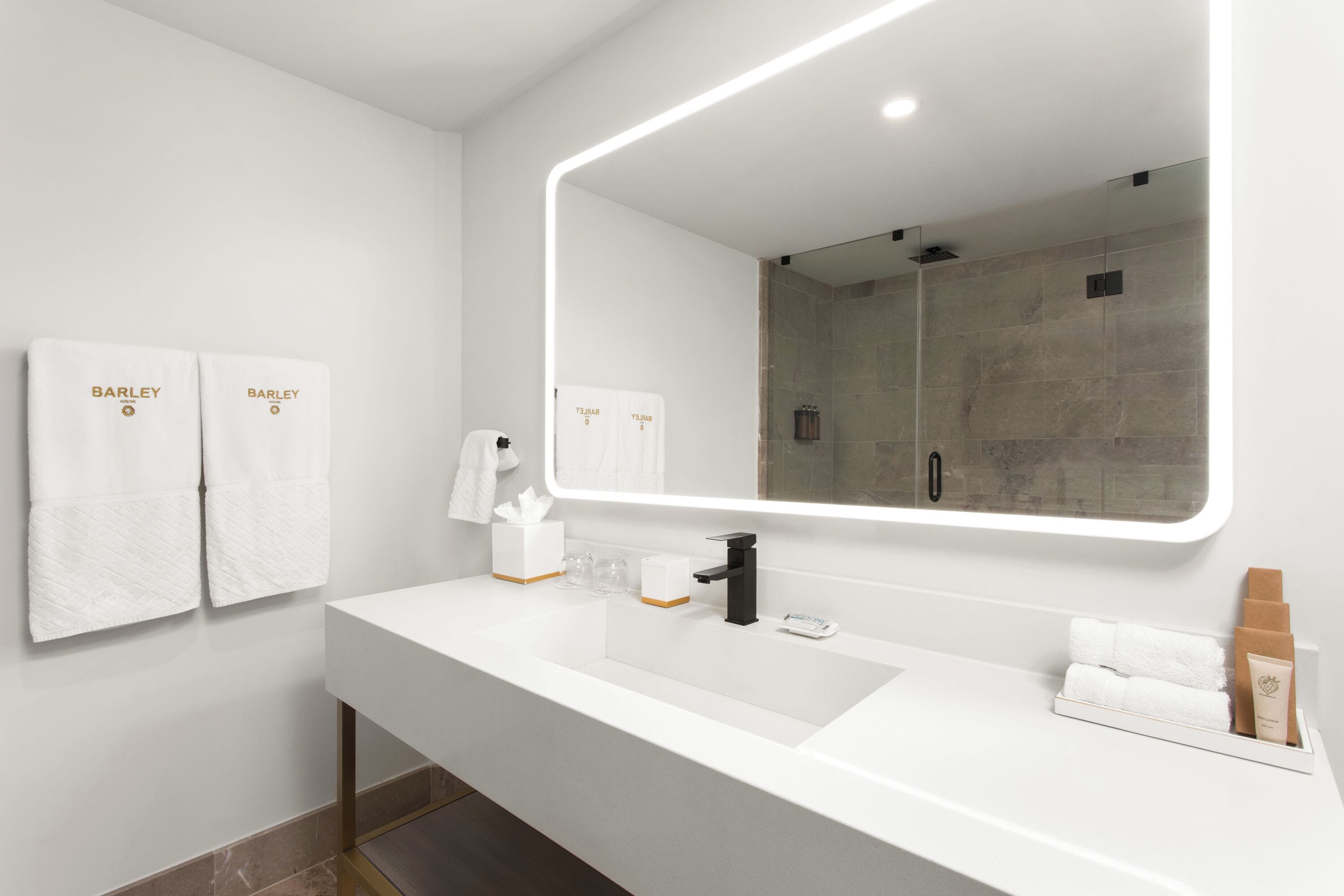 A modern bathroom with a large mirror, two white towels with 'BARLEY' branding, a black faucet, and a white countertop.