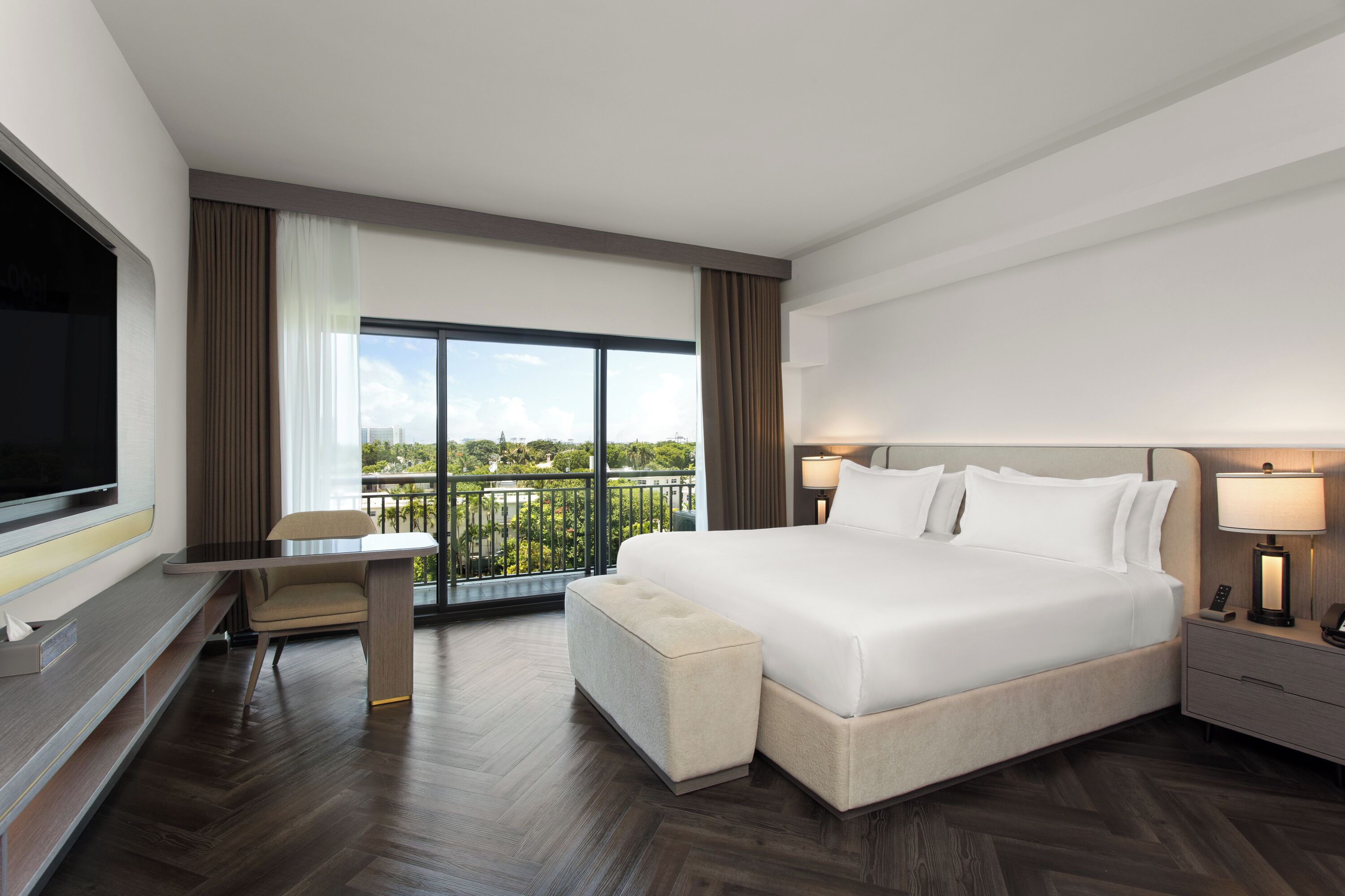 A hotel room with a large bed, a desk with a chair, a TV, and a balcony with a view of the city.