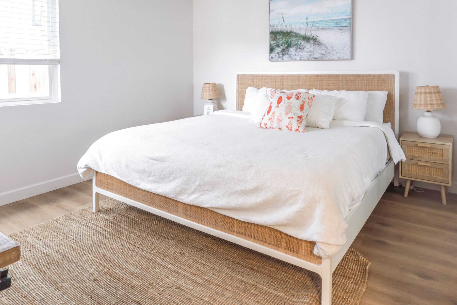 A bedroom with a bed, two bedside tables, a nightstand, a window, and a beach-themed painting on the wall.