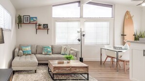 Un salon moderne comprenant un canapé, une table basse et un coin repas. On y trouve de grandes fenêtres, une planche de surf et divers éléments de décoration murale.