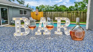 Decoración para fiesta en el jardín con letras iluminadas que dicen 'BAR', dos copas de vino rosado y una botella de vino rosado sobre un suelo de grava.