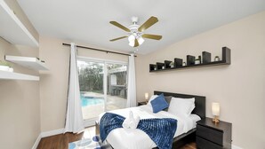 Un dormitorio con cama, mesita de noche, ventilador de techo, ventana con vistas a una piscina y estanterías empotradas.
