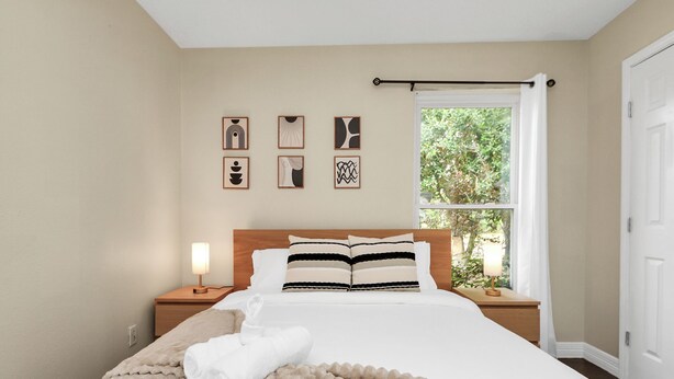 Un dormitorio con cama, una mesita de noche, una lámpara, una ventana con cortinas y decoración en la pared.