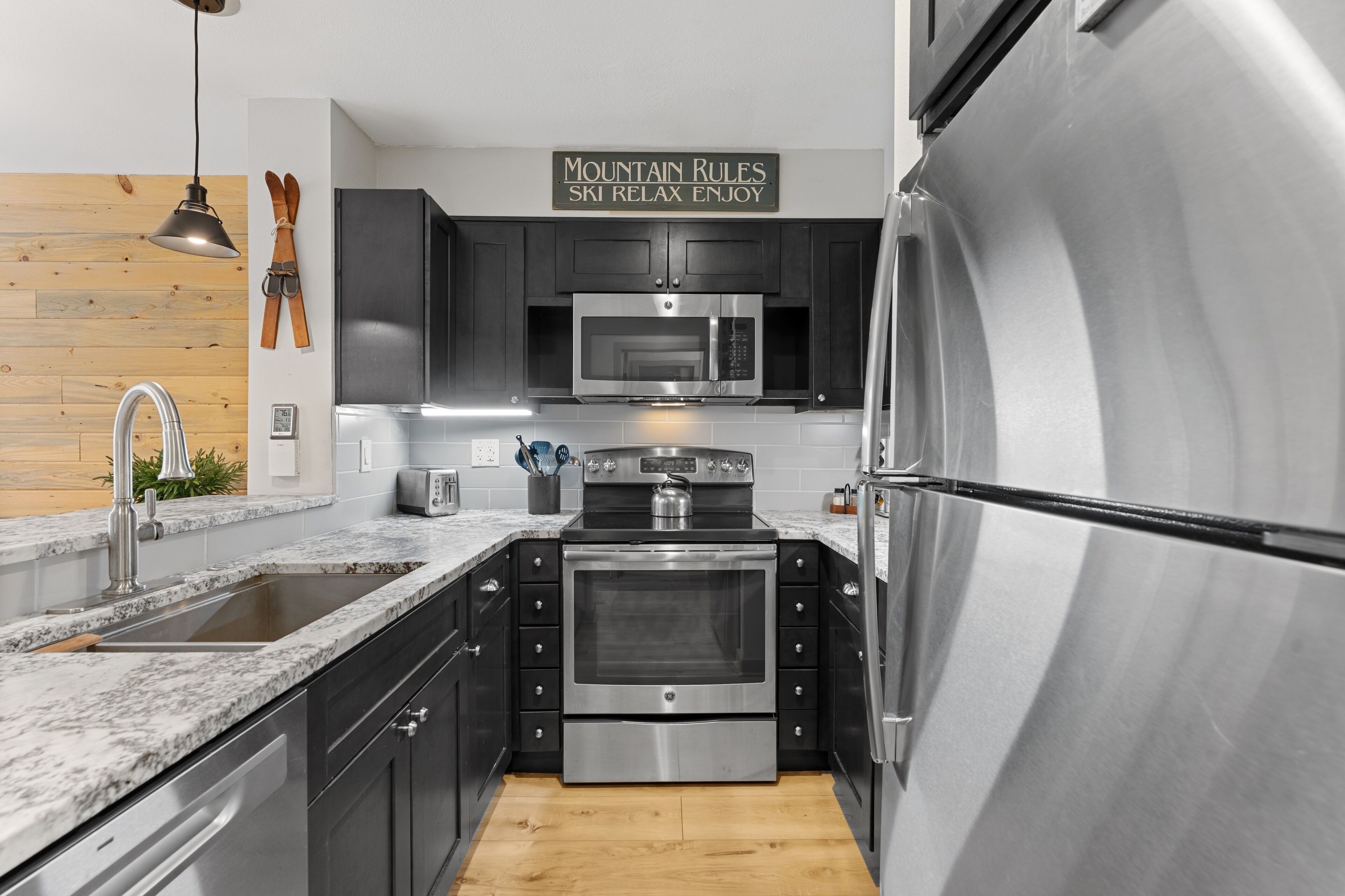 A modern kitchen with stainless steel appliances, wooden flooring, and a sign that reads 'MOUNTAIN RULES SKI RELAX ENJOY'.