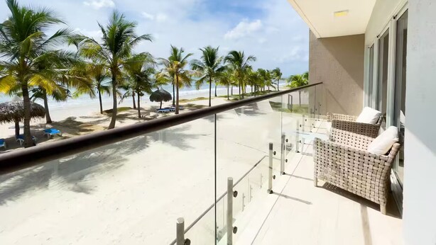 Ein Balkon mit Glasgeländer mit Blick auf einen Strand, Palmen und einen klaren Himmel.