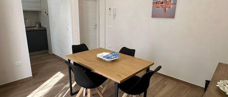 A modern dining area with a wooden table and black chairs, a small kitchen in the background, and a cityscape painting on the wall.
