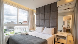 A modern hotel room with a large bed, a bedside table, a desk, and a large window offering a city view. - Condominium With Pool in Downtown Florianopolis - Cjos (Florianópolis)