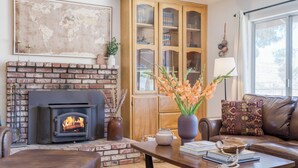 A cozy living room with a brick fireplace, a wooden cabinet, a leather sofa, and a coffee table with books and decorative items. - Desert Shadows by Avantstay Breathtaking Views & Pool (Joshua Tree)
