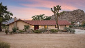A stucco house with a red-tiled roof, a red door, and a small front yard with cacti and rocks. - Desert Shadows by Avantstay Breathtaking Views & Pool (Joshua Tree)
