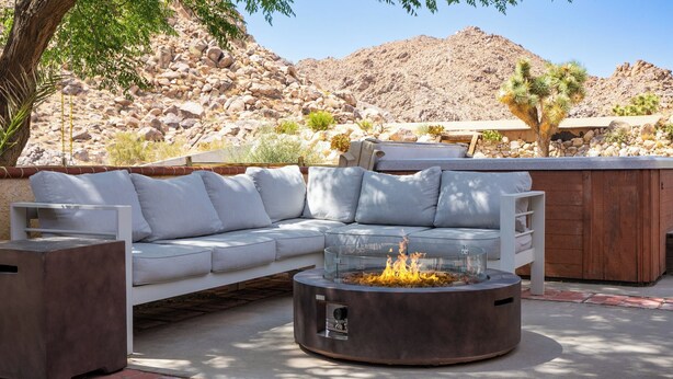 A modern outdoor seating area with a stone fire pit, a white sofa, and a wooden table. - Desert Shadows by Avantstay Breathtaking Views & Pool (Joshua Tree)