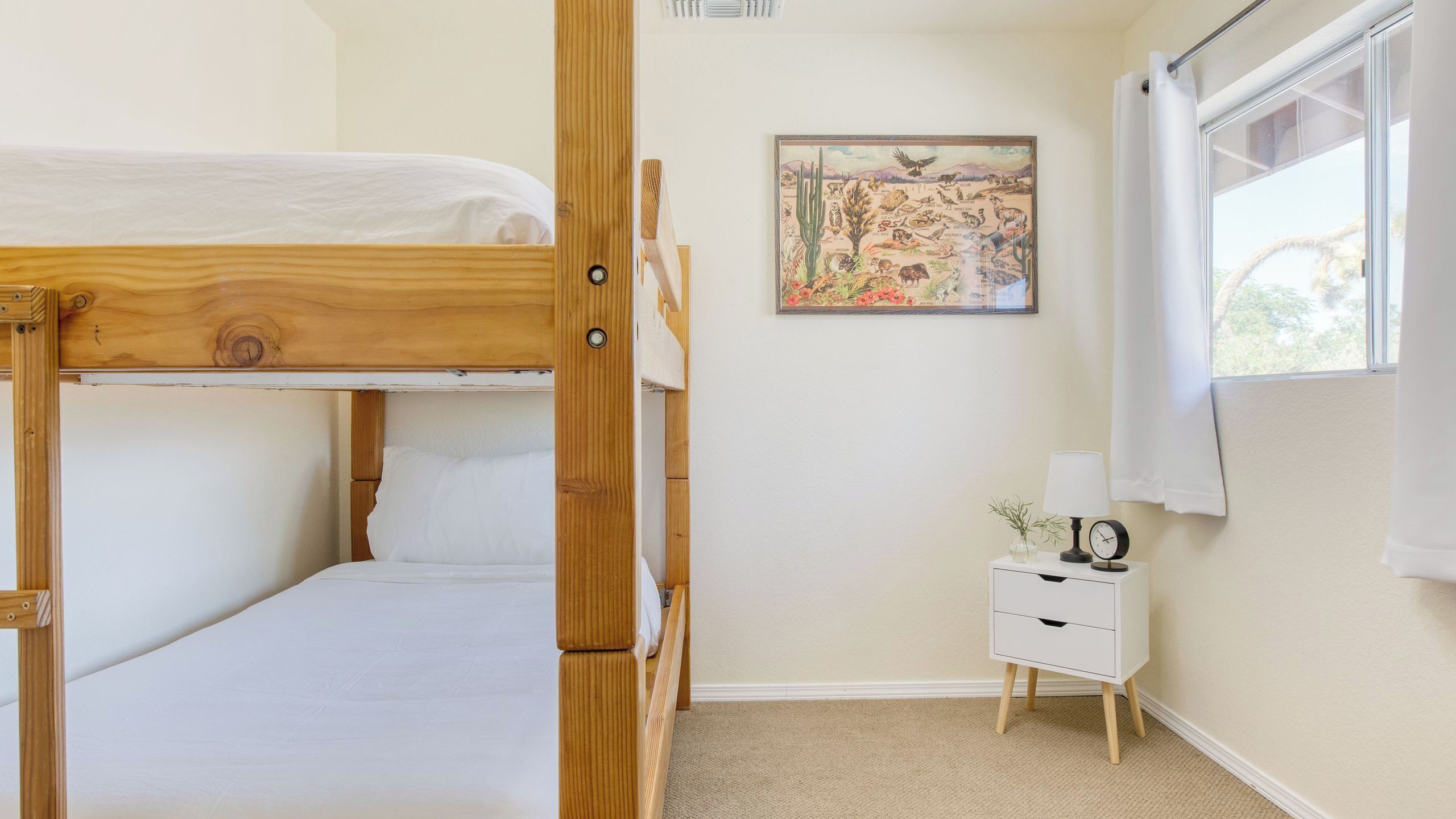 Une chambre avec un lit superposé, une petite table de chevet, un tableau accroché au mur et une fenêtre avec des rideaux.