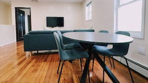 Ein geräumiges Zimmer mit Holzboden, einem runden Tisch mit Stühlen, einer Couch und einem an der Wand befestigten Fernseher.